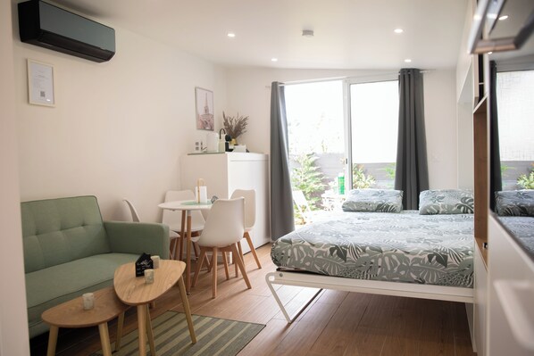 Ein modernes Zimmer mit einem grünen Sofa, einem kleinen Tisch, einem Bett mit gemusterten Bettbezügen, einem Schreibtisch mit Stühlen und einem Fenster mit Vorhängen.