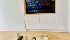 Una sala de estar moderna con un televisor de pantalla plana montado en una pared blanca, una mesa de madera con controles remotos y un vaso de agua.