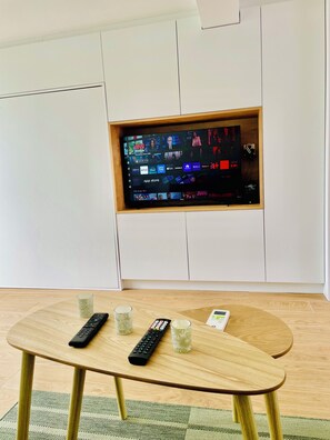 Ein modernes Wohnzimmer mit einem an einer weißen Wand befestigten Flachbildfernseher, einem Holztisch mit Fernbedienungen und einem Glas Wasser.
