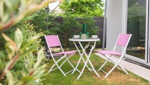 Un patio al aire libre con dos sillas blancas plegables y una pequeña mesa redonda, rodeado de follaje y un ventanal de vidrio.