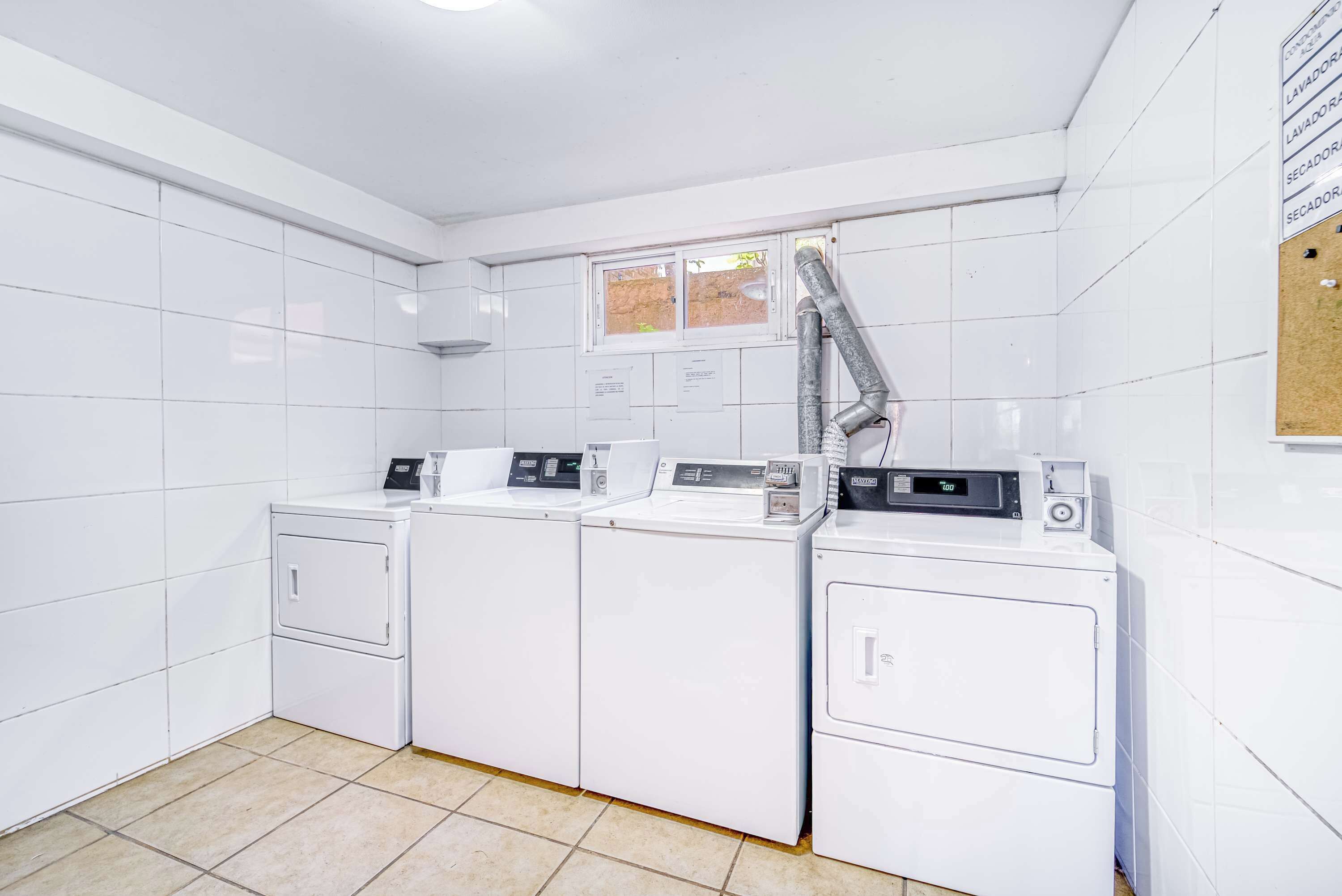 A laundry room with four washing machines and a dryer, white tiled walls, a window, and a bulletin board.
