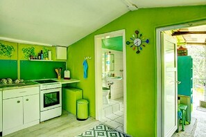 Una cocina compacta con gabinetes blancos, un lavamanos y una estufa. Se observa un cubo de basura, y un reloj está colgado en la pared.