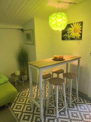 Una habitación pequeña y acogedora con una mesa de madera, tres taburetes, un cojín verde, una alfombra estampada, una lámpara verde colgante y un cuadro de un girasol en la pared.