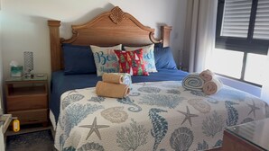 Un dormitorio con cabecera de madera, cama con ropa de cama azul, almohadas decorativas, mesita de noche con lámpara y ventana con persianas.