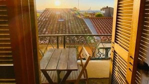 Un balcon avec une table et des chaises en bois, une vue sur les toits et un ciel dégagé.