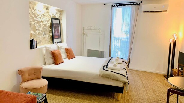 A hotel room with a bed, a chair, a small table, a window with curtains, and an air conditioning unit. - "Ribot" Coquet studio in the heart of Sète, Air conditioning & Wifi (Sète)