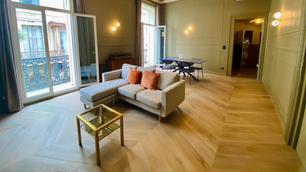 Amplia sala de estar con un sofá grande, una mesa de centro de vidrio y una mesa de comedor con sillas. Hay ventanas grandes que ofrecen luz natural y vistas al exterior.
