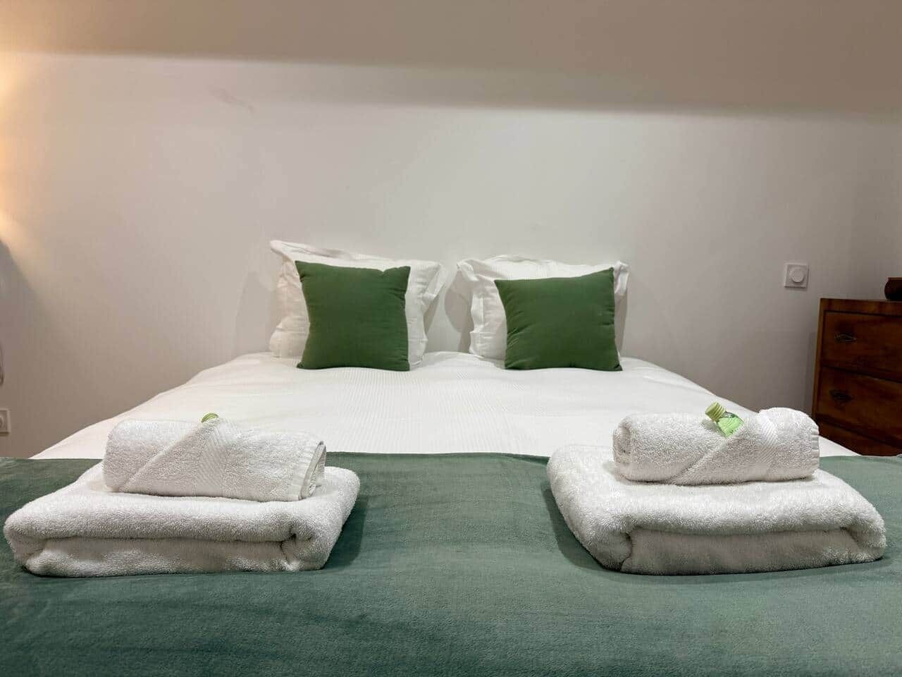 A neatly made bed with white linens, two green pillows, and two folded towels on a green bedspread.