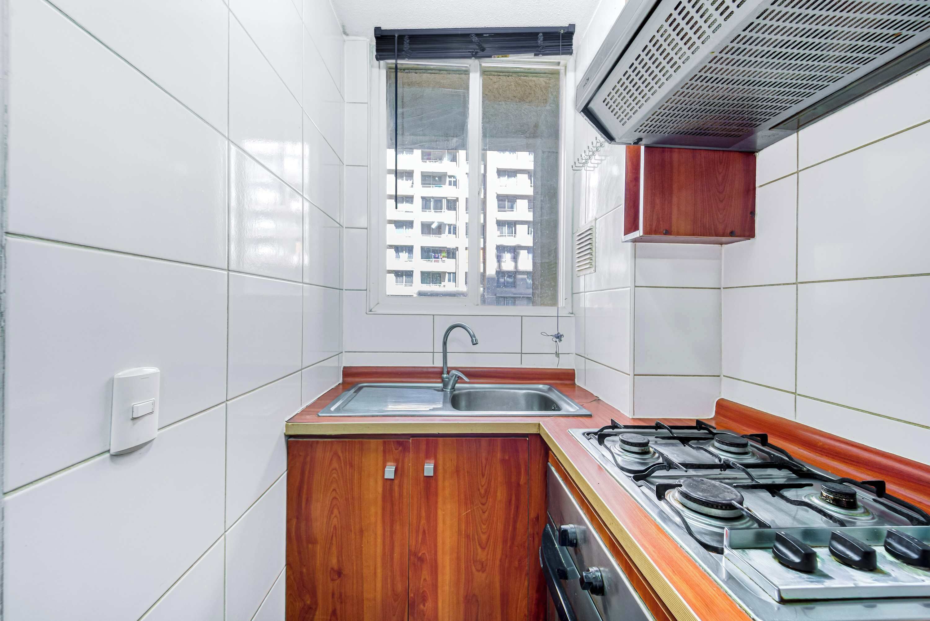 Cocina compacta con azulejos blancos, un mueble de madera, una estufa a gas y una ventana con vistas a edificios.
