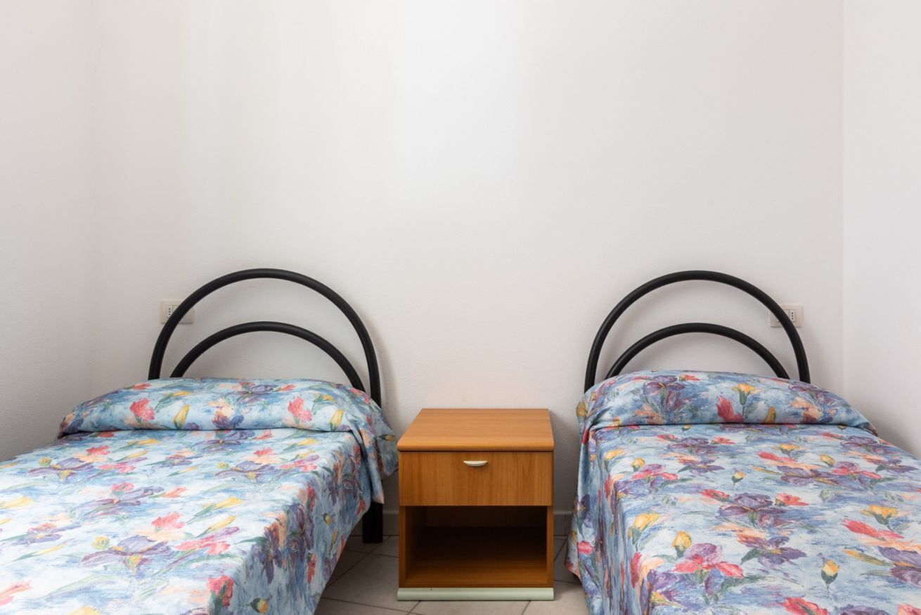 Ein kleines Zimmer mit zwei Einzelbetten, jeweils mit einem blumigen Bettbezug, einem dazwischenliegenden Nachttisch aus Holz und einer schlichten Wand.