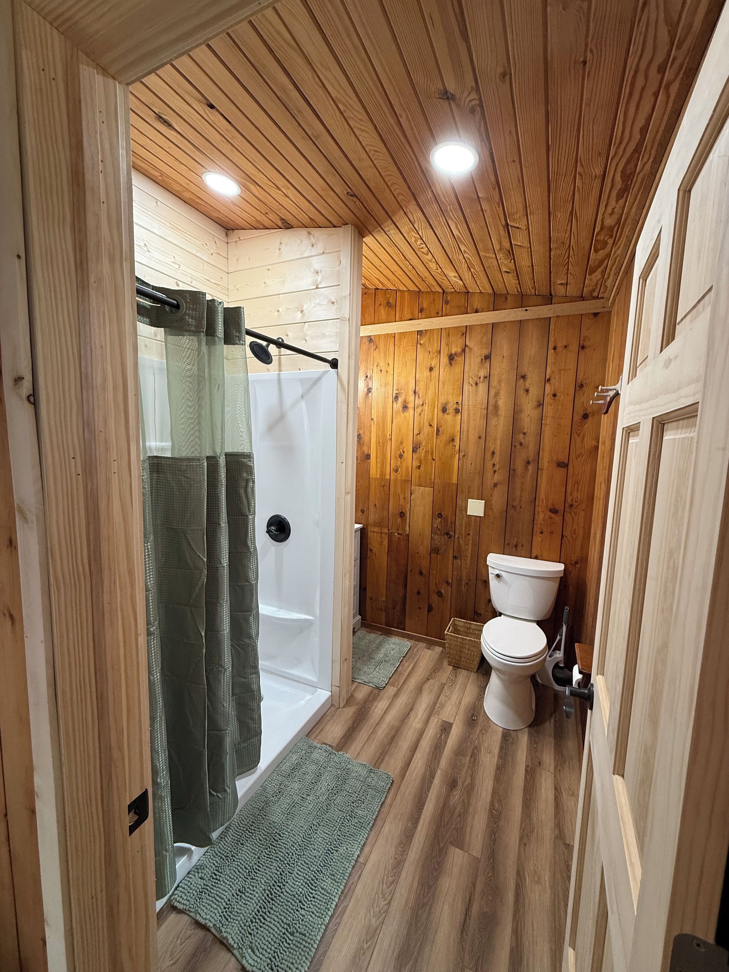 A wooden-paneled bathroom with a shower, toilet, and a green rug on the floor.