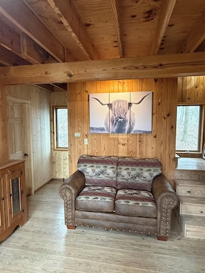 A wooden cabin interior with a leather sofa, wooden walls, and a large window.
