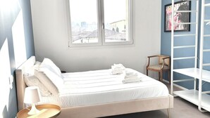 Ett sovrum med en säng, ett nattduksbord med en lampa, en stol, en garderob och ett fönster med utsikt över byggnader.