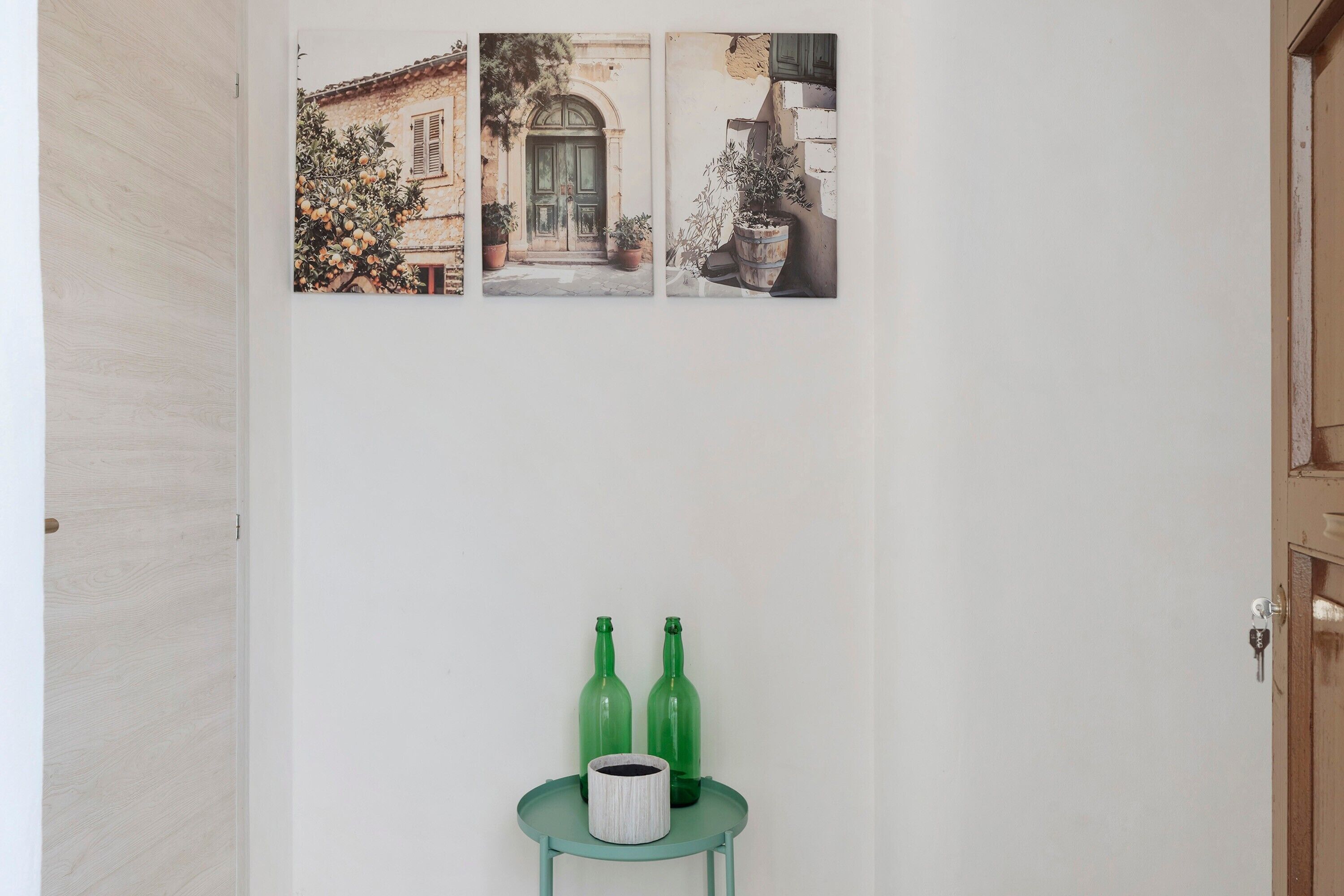 Un couloir avec une porte, un tableau accroché au mur et une petite table sur laquelle se trouvent deux bouteilles vertes et un récipient blanc.