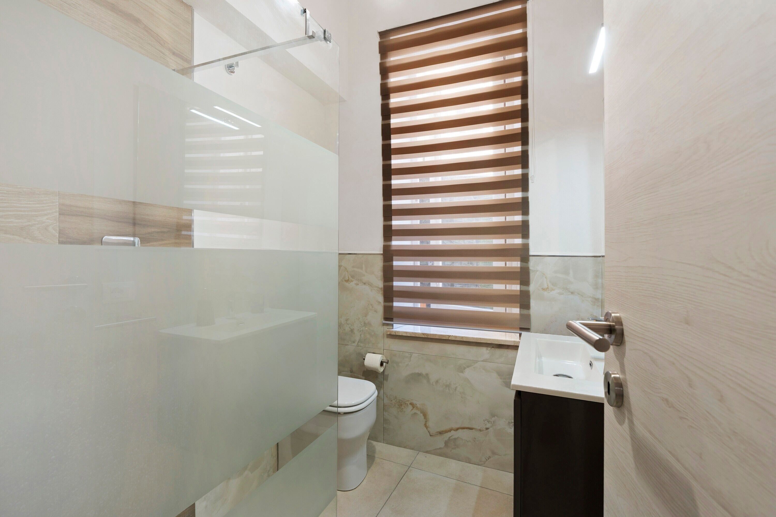 Ein modernes Badezimmer mit Duschkabine aus Glas, Toilette, Waschbecken und einem Fenster mit Jalousien.