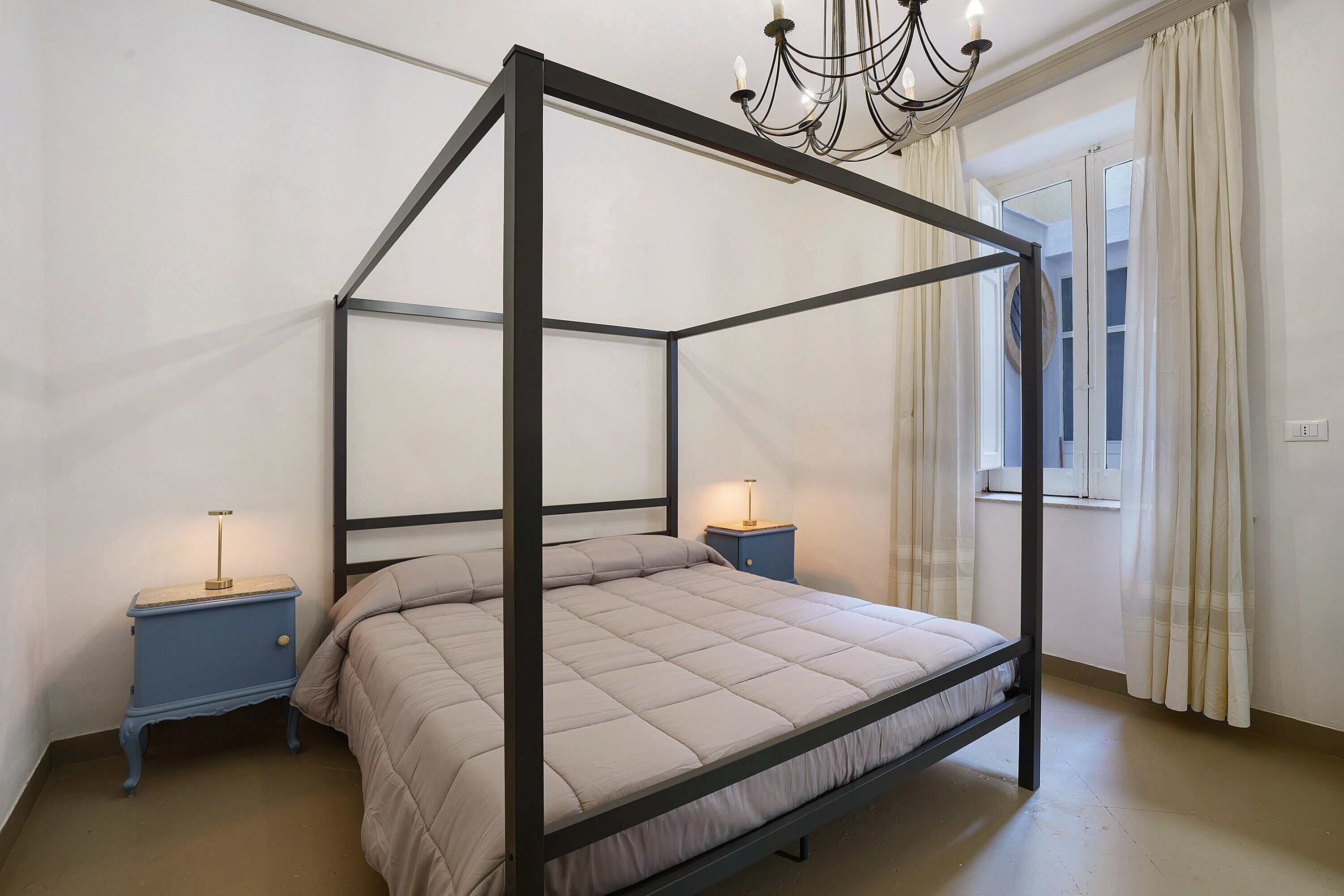 Ein Bett mit Baldachin, ein Kopfteil, zwei Nachttische, eine Chandelüster und ein Fenster mit Vorhängen.