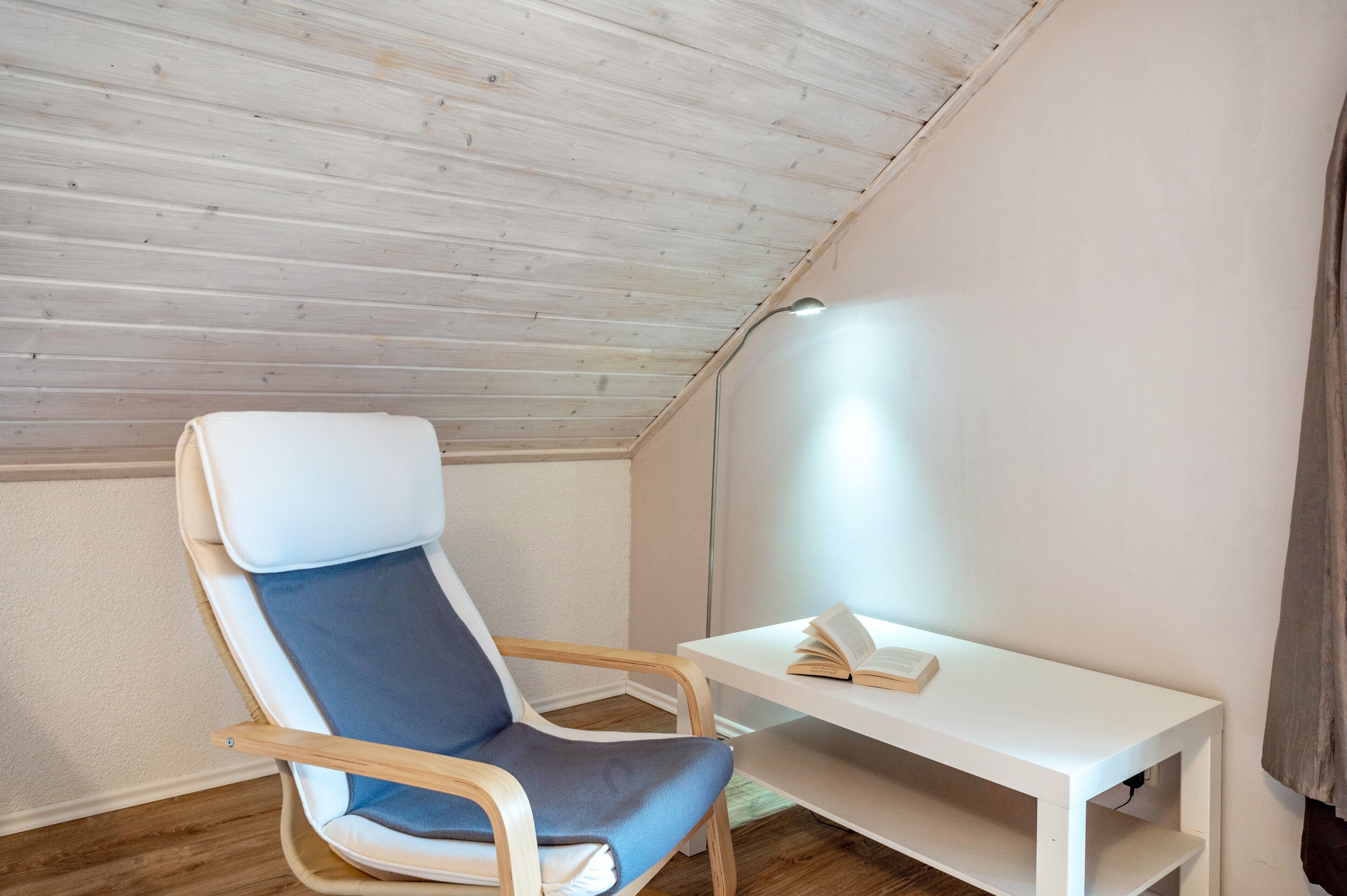 Ein Zimmer mit schrägverdeckter Decke, ein einzelner Sessel mit Kissen, ein kleiner weißer Tisch mit einem Buch und eine Lampe.