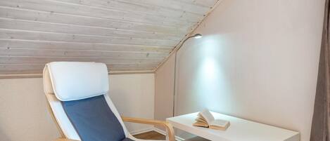 Ein Zimmer mit schrägverdeckter Decke, ein einzelner Sessel mit Kissen, ein kleiner weißer Tisch mit einem Buch und eine Lampe.