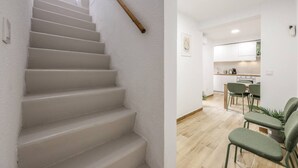Un escalier avec une rampe, un coin cuisine doté de meubles blancs et un espace repas avec des chaises vertes.