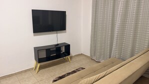 A living room with a TV mounted on the wall, a black entertainment unit, a beige sofa, and a patterned curtain. - Private studio apartment with Smart TV and air conditioning. (Belo Horizonte)