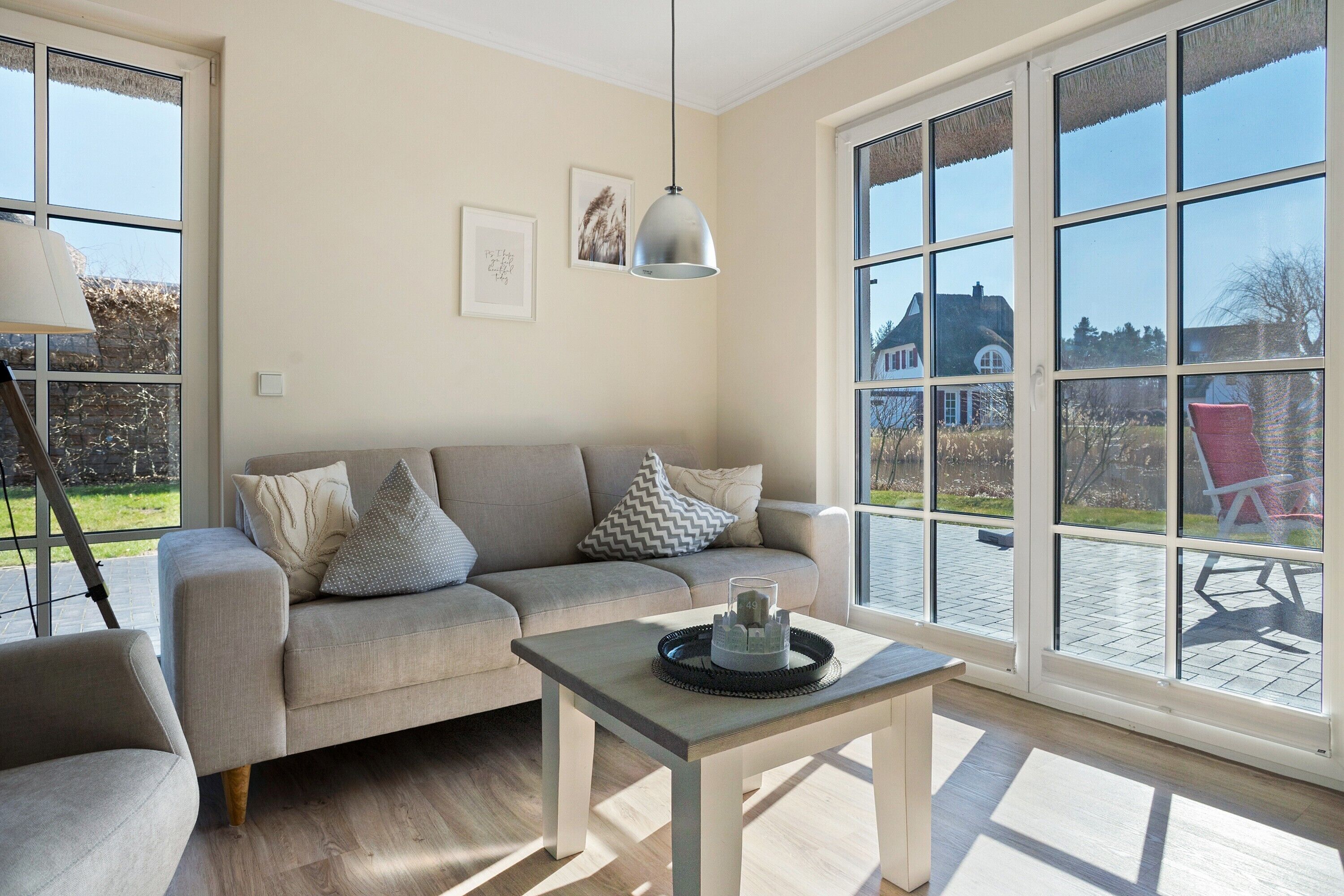 En stue med en sofa, et bord med bakke, en hængende lampe og store vinduer med udsigt til et hus og en have.