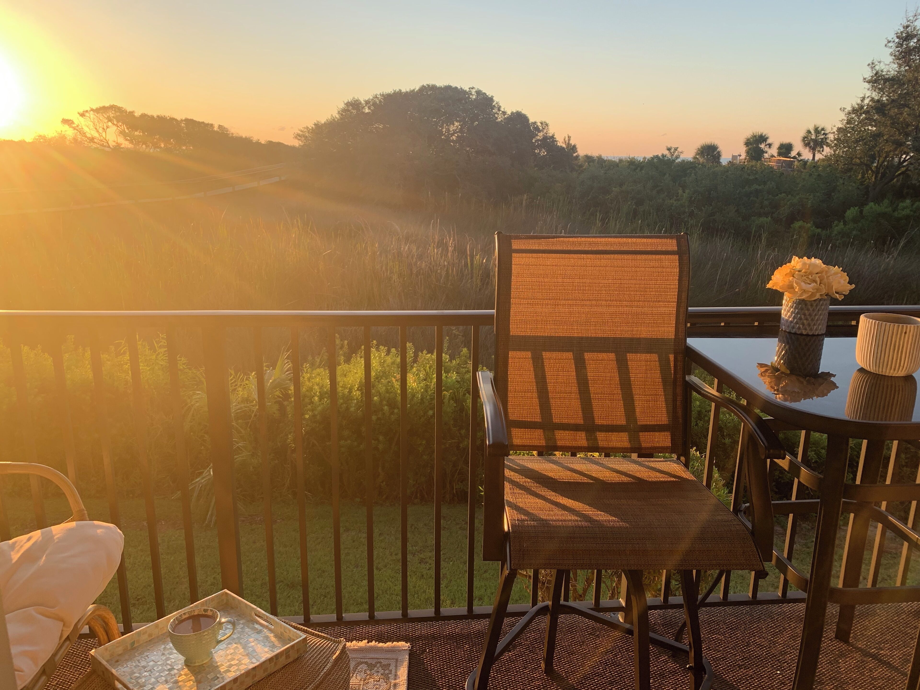 A balcony with a wicker chair, a table with a vase of flowers, and a tray with a cup.