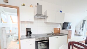 Une cuisine compacte avec des meubles blancs, un four encastré, une machine à café et un téléviseur posé sur une étagère.