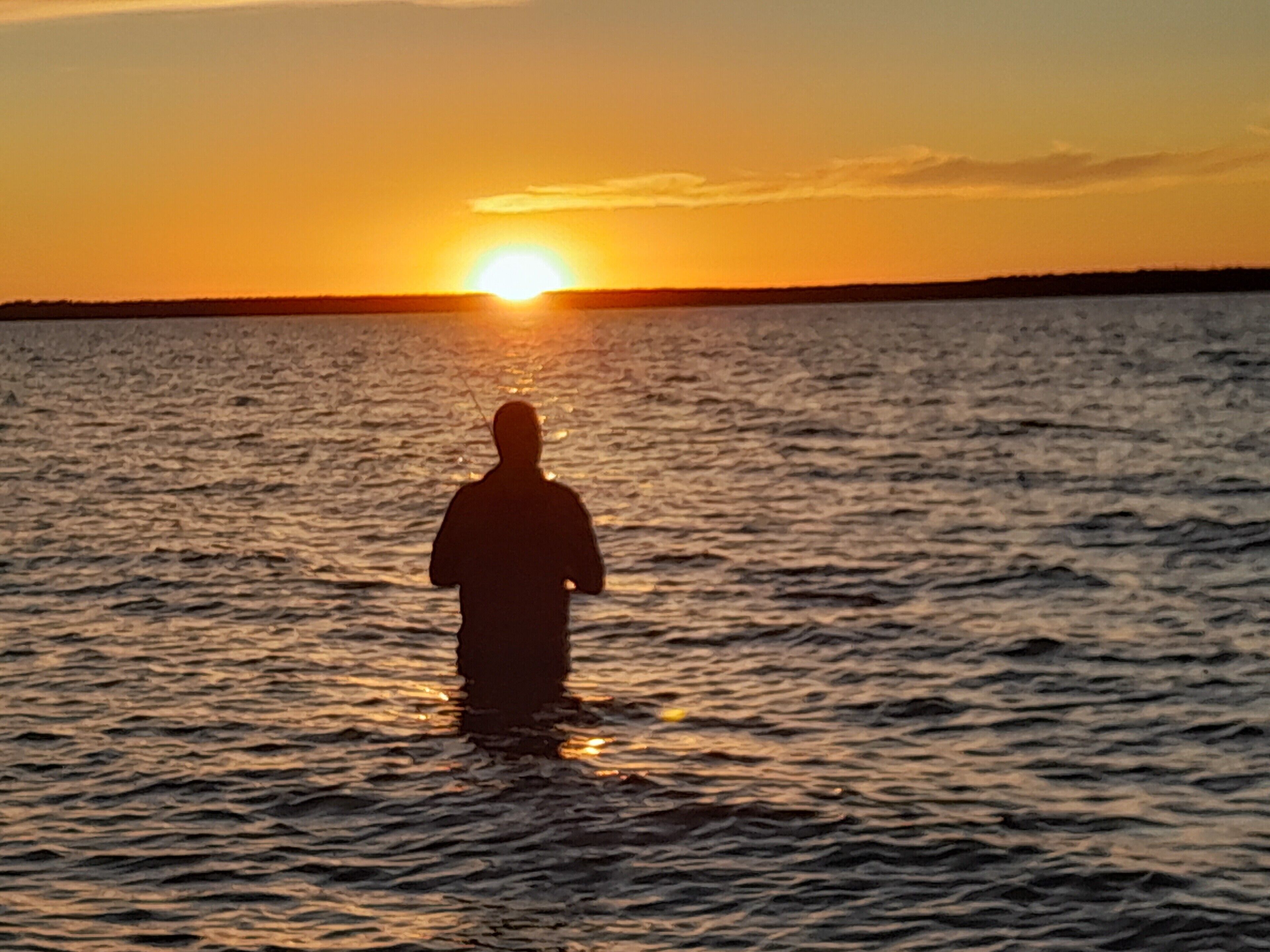 Eine Silhouette einer Person, stehend im Wasser, mit untergehender Sonne im Hintergrund.