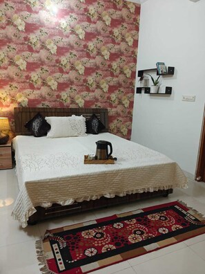 A bedroom with a floral wallpaper, a bed with a white quilt, a bedside table with a lamp, and a decorative rug. - Modern Family Stay with Garden (Jaipur)
