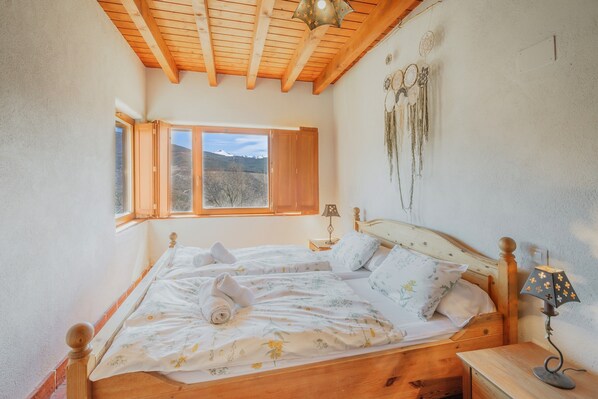 Un dormitorio con una cama de madera, una mesita de noche, una lámpara, un ventanal con vistas al exterior y una pared decorada con atrapasueños.