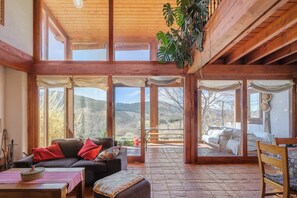 Una habitación amplia con techo de madera, ventanas grandes, un sofá con cojines, una mesa y una planta colgante.