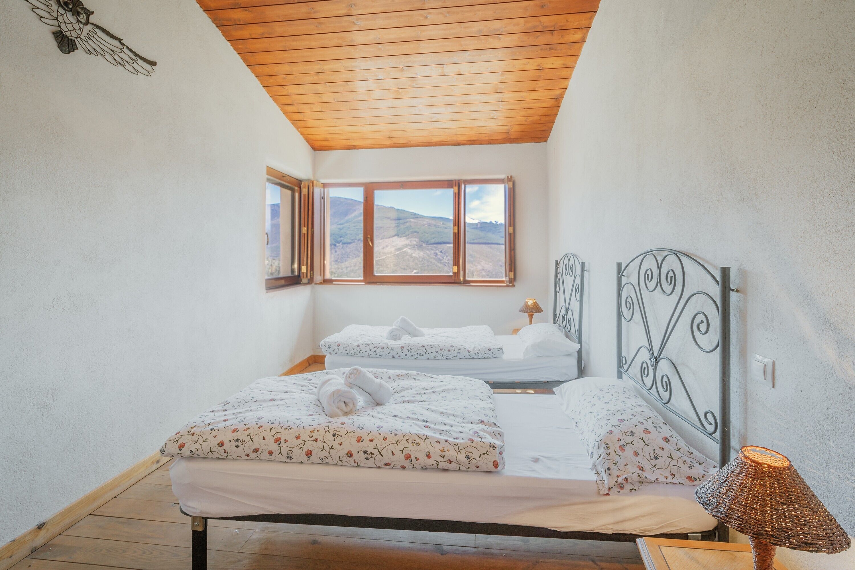 Une chambre avec un plafond en bois, un lit recouvert d’une literie à motifs floraux, une table de chevet avec une lampe, et une vue sur l’extérieur par une fenêtre.