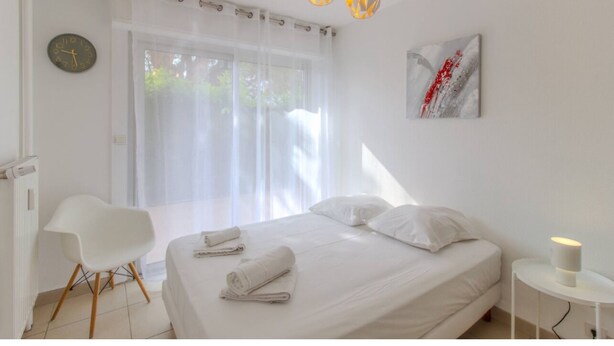 Quarto moderno com cama, cadeira, mesa de cabeceira e janela com cortinas leves.