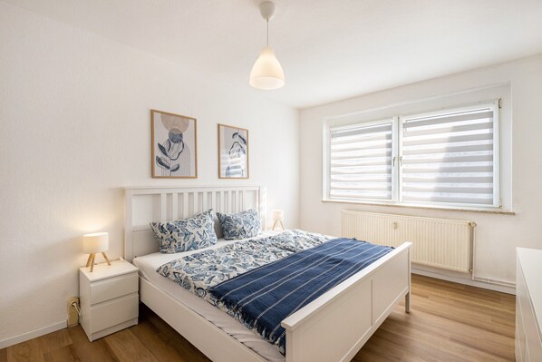 Ein Schlafzimmer mit Bett, Nachttischen, einer Pendelleuchte, einem Fenster mit Jalousien und Wandbildern.