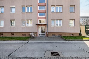 Ein mehrstöckiges Gebäude mit einem zentralen Eingang, einer kleinen Außenbereich-Sitzgelegenheit und einem gepflasterten Vorplatz.