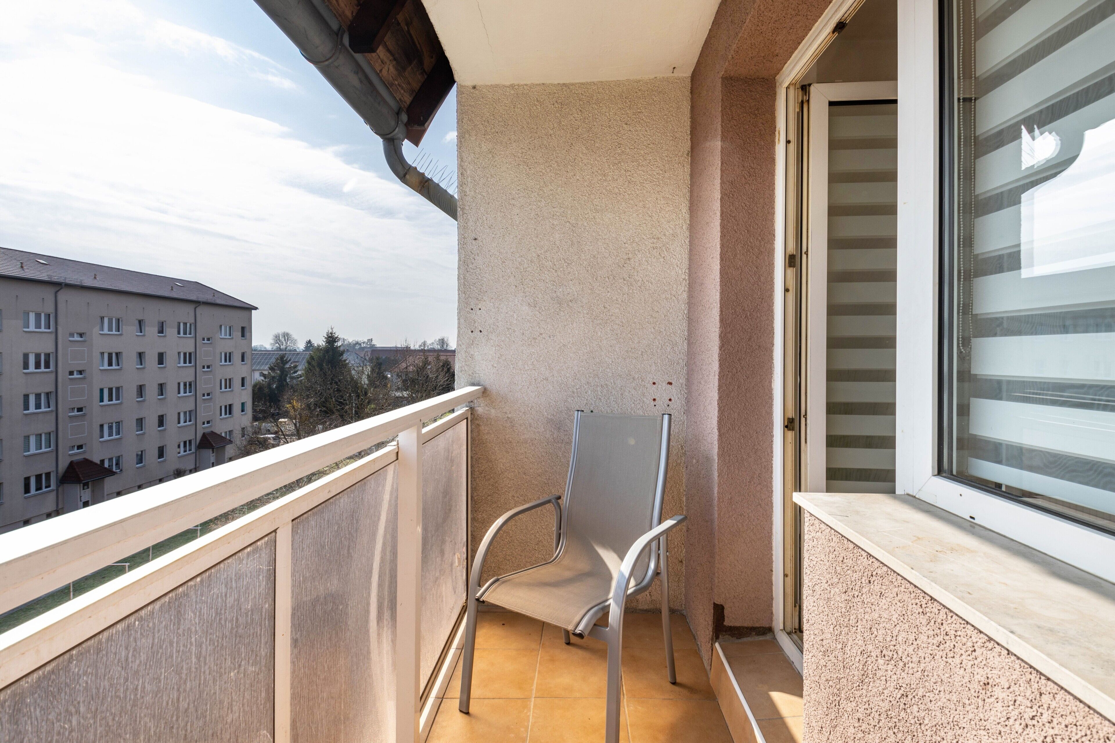 A balcony with a metal chair, a window with blinds, and a view of buildings.