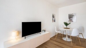 Una sala de estar moderna con un televisor de pantalla plana sobre una consola blanca, una mesa de comedor con sillas blancas y una pequeña planta en maceta.