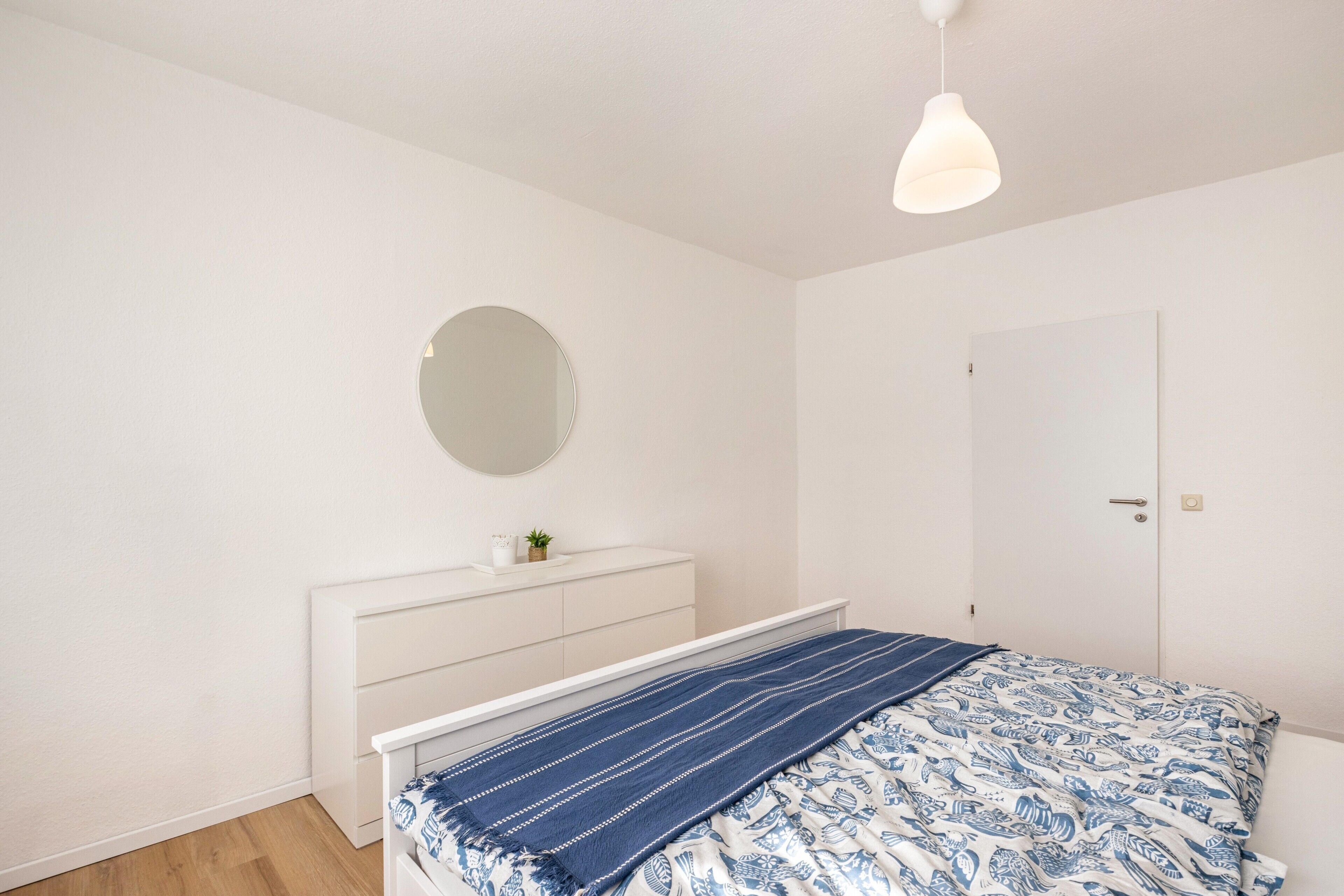 Un dormitorio con cama, mesita de noche con espejo, armario blanco y lámpara colgante.