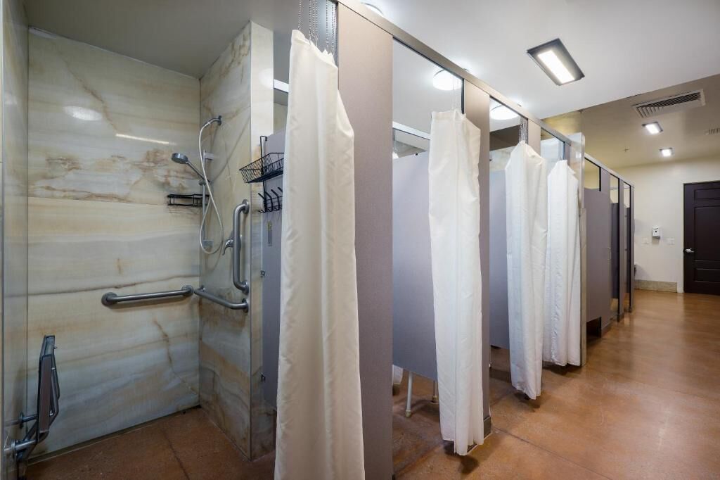 A row of white shower stalls with glass doors, a handrail, and a marble wall.