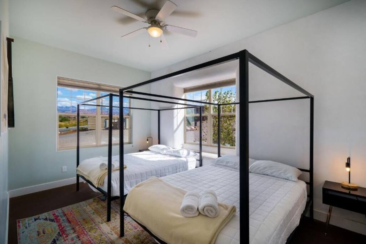 A four-poster bed room with two beds, a ceiling fan, and large windows offering a view of the outdoors.