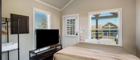 A bedroom with a bed, a TV on a stand, a wardrobe, and two windows with blinds.