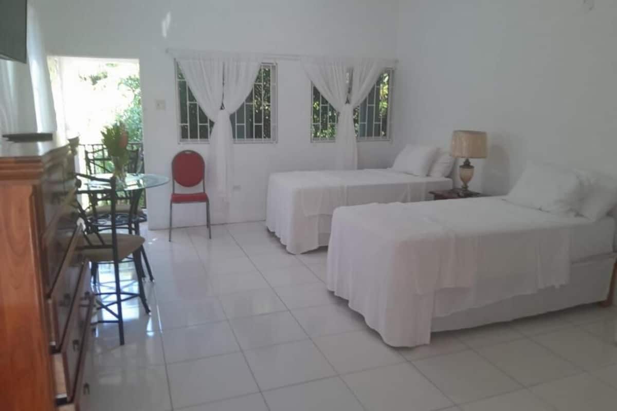 Una habitación con dos camas, una mesa de comedor con sillas, una mesita con lámpara y un ventanal con cortinas.