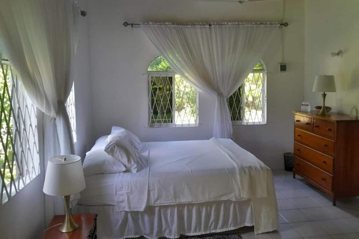 Un dormitorio con una cama, una mesita de noche, un aparador y dos ventanas con cortinas.