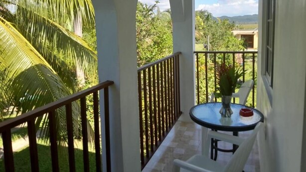 A balcony with a table and chairs, surrounded by a white railing, overlooking a lush green landscape with palm trees and a clear sky. - Coco Gardens (Negril)