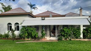 A single-story house with a white exterior and a red-tiled roof. There is a covered porch with a lattice design and a small step leading to the entrance. The house is surrounded by a well-maintained lawn and various plants. - Coco Gardens (Negril)
