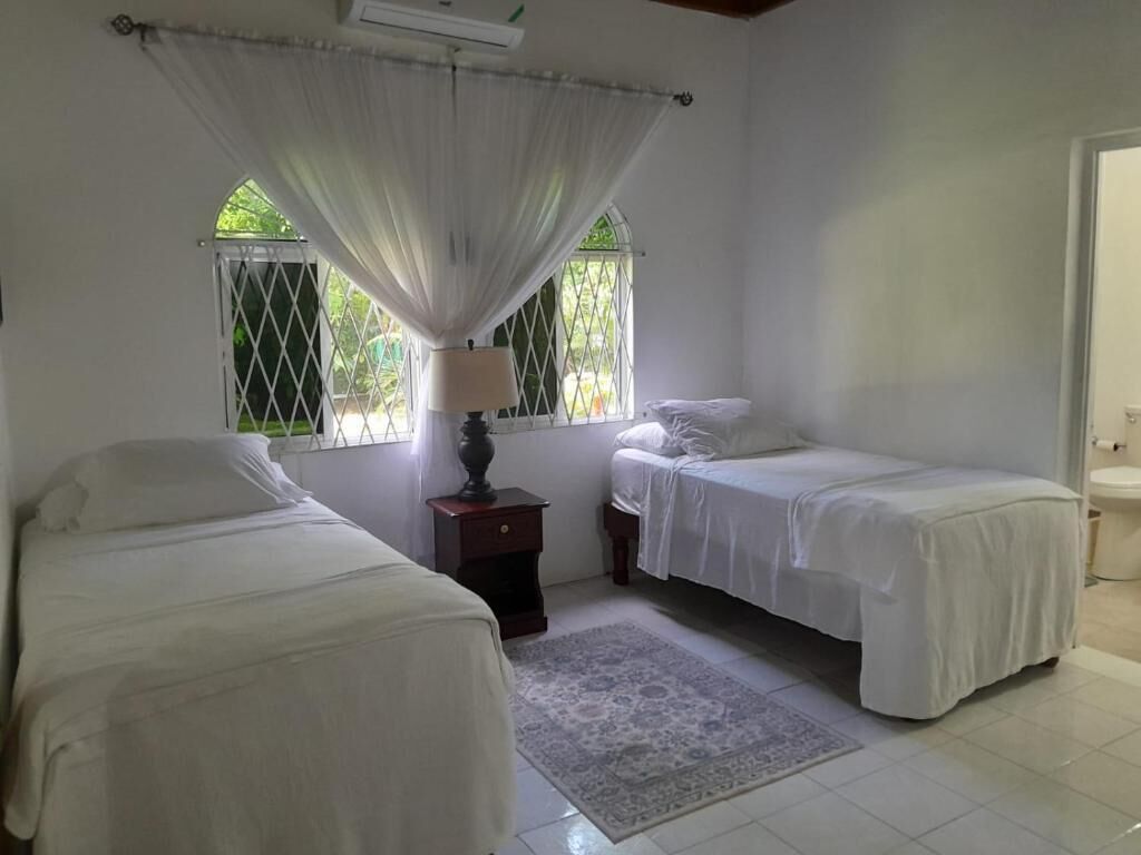 A room with two single beds, a nightstand with a lamp, a window with a view of greenery, and a door leading to a bathroom.