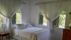 A bedroom with a bed, curtains, a nightstand, a lamp, and a dresser. - Coco Gardens (Negril)
