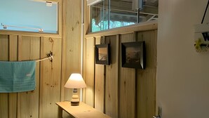 A wooden interior with a window, a towel hanging on a hook, a lamp on a wooden table, and two framed pictures on the wall. - Villa dos Sonhos Vá de Caiaque Até o Mar (São Francisco do Sul)