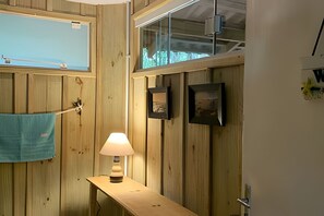 Un intérieur en bois avec une fenêtre, une serviette accrochée à un crochet, une lampe posée sur une table en bois et deux tableaux encadrés au mur.