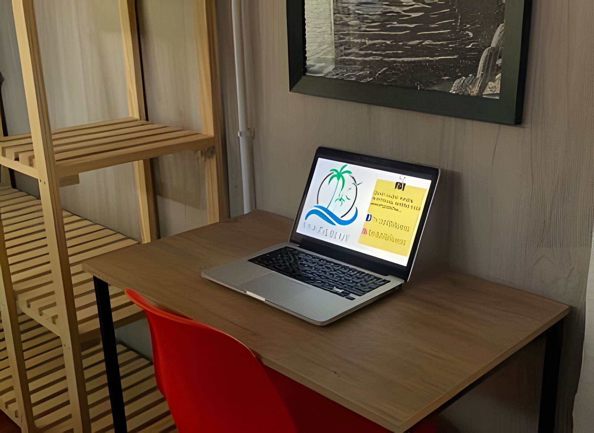 Una computadora portátil sobre una mesa de madera con una silla roja, un cuadro en la pared y una estructura de literas.